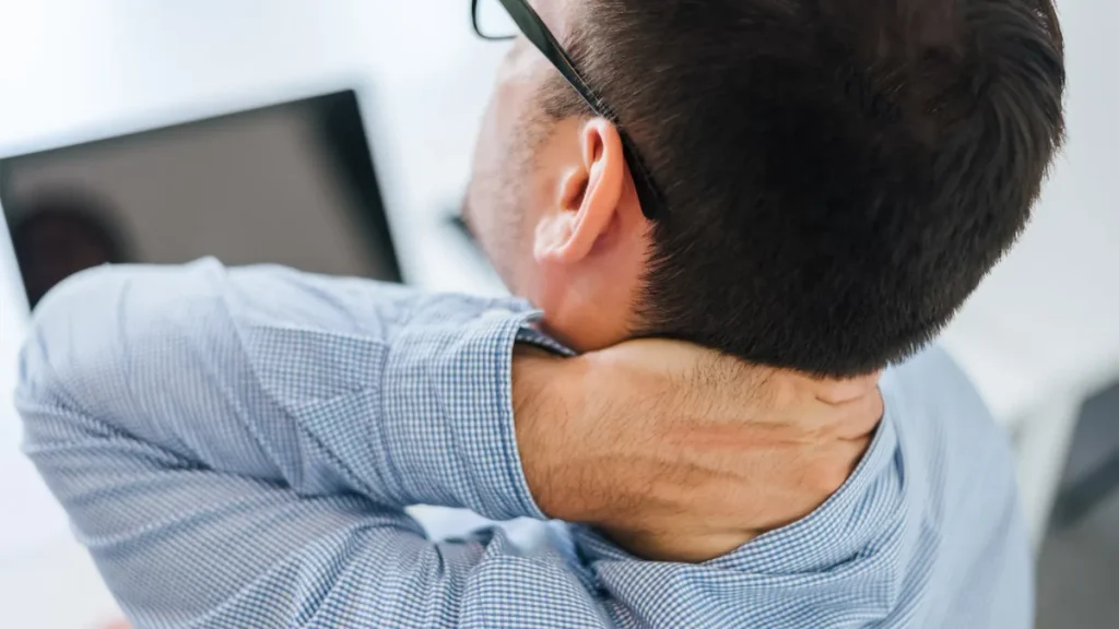 Hombre con gafas se masajea la nuca frente al ordenador por la tensión acumulada en el cuello