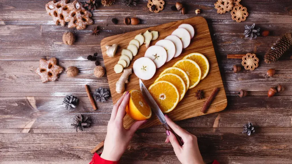 Navidad saludable: manos que cortan naranja, manzana y jengibre sobre una tabla rodeada de adornos navideños